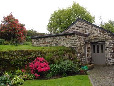 A stone building with a door surrounded by a garden with green grass and flowering plants at Burham Barn in Dousland near Yelverton