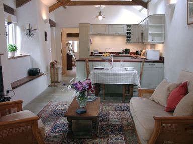 A combined kitchen and living room with a dining table set for two and a coffee table with flowers at Burham Barn in Dousland near Yelverton