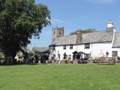 A white building with a slate roof and outdoor seating on a lawn with a stone tower in the background at Burham Barn in Dousland near Yelverton