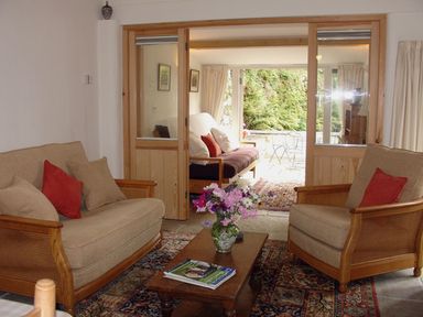 A living room with sofas and a coffee table with flowers and magazines separated by wooden framed glass doors from a sunroom with a sofa and outdoor furniture at Burham Barn in Dousland near Yelverton