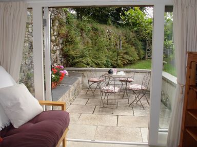 A patio with metal chairs and table with cushions viewed through sliding glass doors from a room with a wooden-framed sofa Burham Barn Dousland near Yelverton
