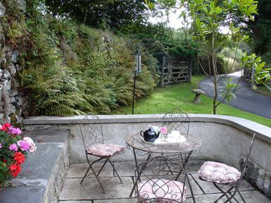 An outdoor patio area with metal table and four chairs with cushions near a garden and stone wall at Burham Barn in Dousland near Yelverton