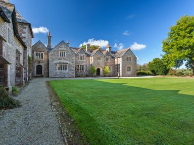 A large stone manor house with multiple chimneys and leaded windows facing a gravel path and a lawn with trees at Great Bidlake Manor in Bridestowe near Lydford