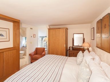 A bedroom with a bed with pillows a wooden wardrobe a dressing table with a mirror and lamp an armchair and an open door to a bathroom at Great Bidlake Manor in Bridestowe near Lydford