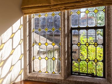 A window with yellow stained glass panes and stone frame with an outside view of rooftops and greenery at Great Bidlake Manor in Bridestowe near Lydford