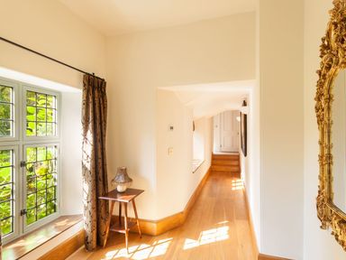 A hallway with wooden floor a window with curtains a small table with lamp and an ornate gold mirror at Great Bidlake Manor Bridestowe near Lydford