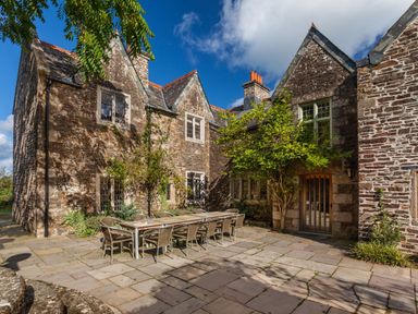 An outdoor patio with a long dining table and chairs in front of a stone house with multiple windows and a door at Great Bidlake Manor in Bridestowe near Lydford