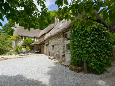 A stone cottage with a thatched roof and outdoor metal table and chairs surrounded by greenery at Bagtor Mill in Ilsington near Haytor Vale