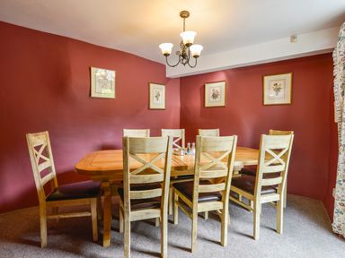 A dining room with a wooden table and eight chairs on carpet with framed pictures on red walls at Bagtor Mill in Ilsington near Haytor Vale