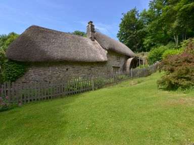 A stone building with a thatched roof and water wheel surrounded by grass and trees at Bagtor Mill Ilsington near Haytor Vale