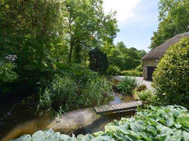 A garden with a small wooden bridge over a pond and a thatched roof building at Bagtor Mill in Ilsington near Haytor Vale
