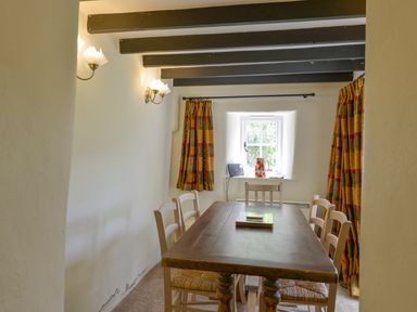 A dining room with a wooden table and six chairs by a window with patterned curtains at Little Gate Cottage in North Bovey