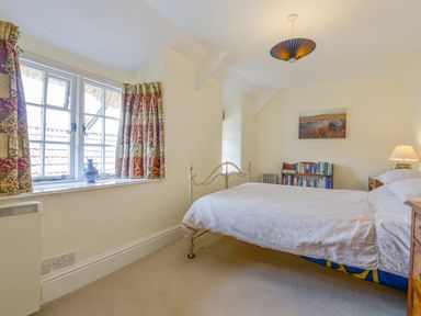 A bedroom with a bed covered in white bedding a window with patterned curtains a bookshelf and a bedside table with a lamp at Little Gate Cottage in North Bovey