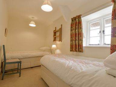 A bedroom with two single beds a wooden chair a bedside lamp a small wooden shelf and a window with patterned curtains at Little Gate Cottage in North Bovey