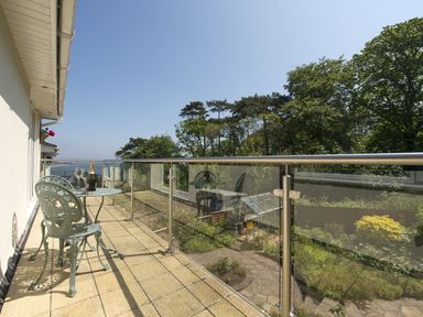 A balcony with a metal table and chairs and a bottle with glasses overlooking a garden and trees at Exe View in Dawlish