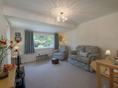 A living room with a patterned sofa and armchair a window with curtains a small table a lamp and a dining table at Exe View in Dawlish