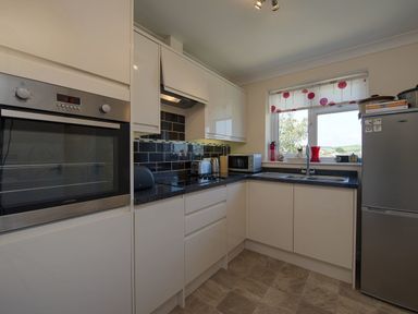A kitchen with white cabinets black countertops an oven a microwave a toaster a kettle a refrigerator and a window with a patterned blind at Exe View in Dawlish