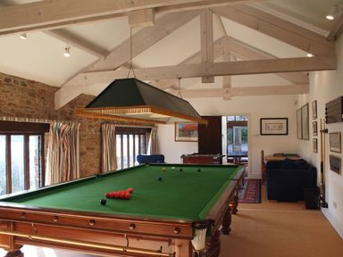 A snooker table in a room with exposed wooden beams windows with striped curtains and seating area at Sandridge Barton in Stoke Gabriel