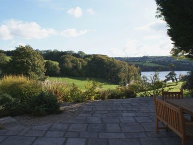 An outdoor patio with wooden chairs and table overlooking a grassy field river and wooded hills at Sandridge Barton in Stoke Gabriel