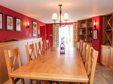 A dining room with a long wooden table surrounded by wooden chairs and a bookshelf against a red wall at Sandridge Barton in Stoke Gabriel