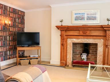 A living room with a wooden fireplace a flat screen tv on a wooden stand and wallpaper with book designs at Sandridge Barton in Stoke Gabriel