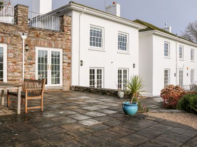An outdoor patio area with stone tiles wooden bench table potted plants and a white house with multiple windows and doors at Sandridge Barton in Stoke Gabriel