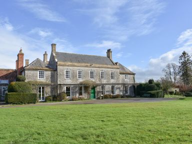 An exterior view of a stone house with multiple chimneys a green door and trimmed hedges in front at Wootton House in Butleigh Wootton near Compton Dundon