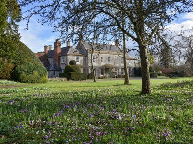 A large stone house with chimneys surrounded by trees and a grassy garden with purple and white flowers at Wootton House in Butleigh Wootton near Compton Dundon