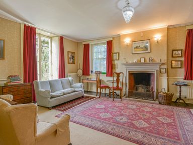 A living room with a fireplace red curtains armchairs a gray sofa and a large patterned rug at Wootton House in Butleigh Wootton near Compton Dundon