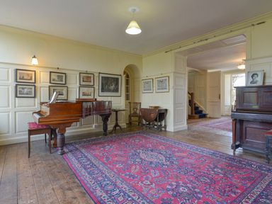 A room with a grand piano a smaller piano framed pictures on paneled walls and a red patterned rug at Wootton House in Butleigh Wootton near Compton Dundon