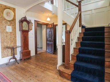 A hallway with wooden floor a staircase with dark carpet a wooden grandfather clock and a small round table at Wootton House in Butleigh Wootton near Compton Dundon