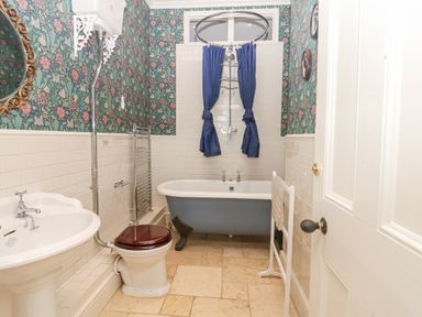 A bathroom with a freestanding clawfoot bathtub with blue curtains a toilet with a wooden seat a pedestal sink and floral wallpaper at Wootton House in Butleigh Wootton near Compton Dundon
