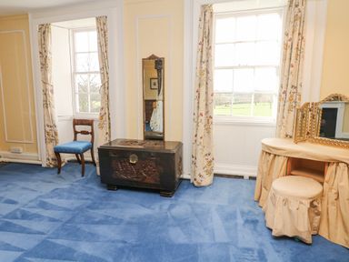 A room with a blue carpet two windows with floral curtains a wooden chair a carved wooden chest a mirror and a covered vanity table with a stool at Wootton House in Butleigh Wootton near Compton Dundon