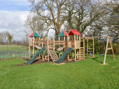 A wooden outdoor playground with slides swings and climbing features on grass at Wootton House in Butleigh Wootton near Compton Dundon