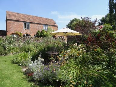 A garden with a table and umbrella surrounded by plants and flowers near a brick house at Pittards Farm Cottage in South Petherton