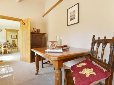 A dining area with a wooden table and chairs including one with a red cushion and a cabinet with decorative items at Pittards Farm Cottage in South Petherton