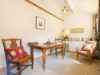 A room with a wooden table and chairs a patterned armchair books on a shelf and a lamp at Pittards Farm Cottage in South Petherton