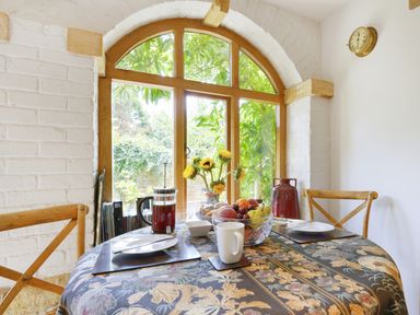 A dining area with a round table covered with a floral tablecloth near arched wooden windows at Pittards Farm Cottage in South Petherton