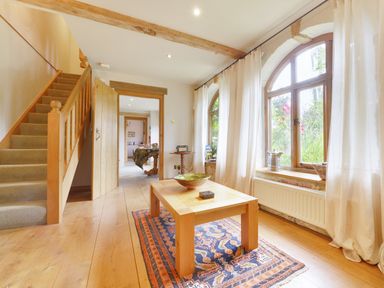 A hallway with wooden stairs a table on a rug and arched windows with curtains at Pittards Farm Cottage in South Petherton