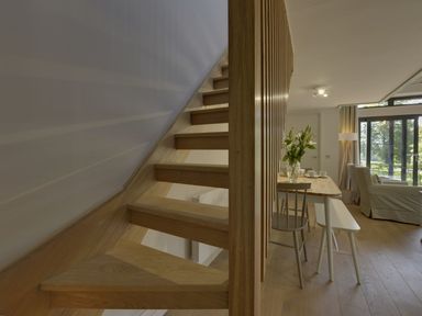A wooden staircase next to a dining table with chairs and a bench and a living area with armchairs and large windows at Grooms Cottage Talaton near Ottery St Mary