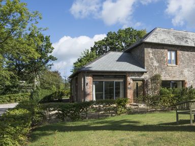 A stone house with a slate roof surrounded by bushes and trees with a wooden bench on the grass at Grooms Cottage Talaton near Ottery St Mary