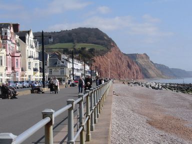 A beach with a promenade railing people walking and sitting buildings and cliffs in the background