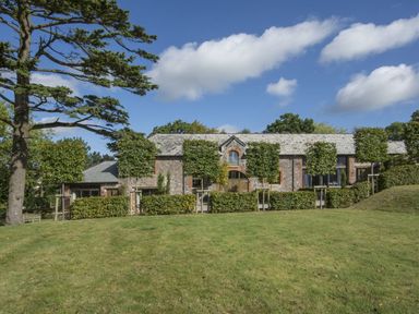 A stone house with a tiled roof behind a row of small trees and hedges on a grassy lawn at Gardeners Cottage in Talaton near Ottery St Mary