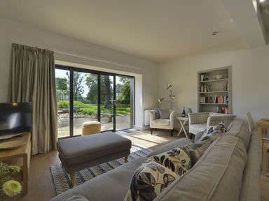 A living room with a sofa, armchairs, bookshelf, ottoman, and sliding glass doors overlooking a garden at Gardeners Cottage in Talaton near Ottery St Mary