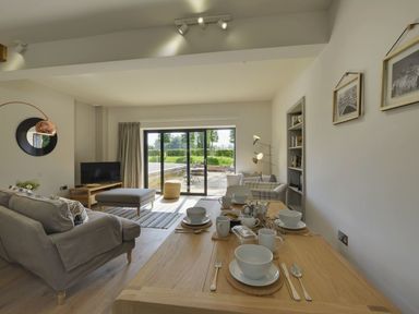 A dining table set with bowls cups and cutlery in a living room with sofas a television and sliding glass doors at Gardeners Cottage in Talaton near Ottery St Mary