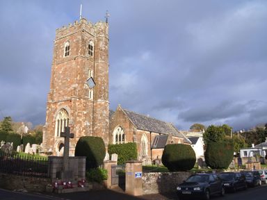 A stone church with a square tower and gravestones in the yard with cars parked along the street at 1 The Quay in Lympstone