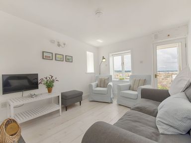A living room with two armchairs a sofa a TV on a white stand and a door to the outside at 1 The Quay in Lympstone