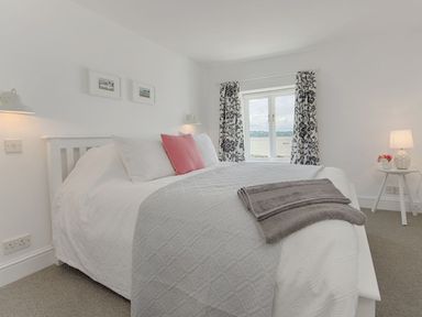 A bedroom with a white bed with white and pink pillows a window with patterned curtains and a small round table with a lamp and flowers at 1 The Quay in Lympstone