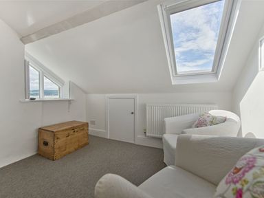 An attic room with two white armchairs with floral cushions a wooden chest a small door a radiator and windows at 1 The Quay in Lympstone