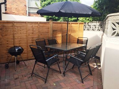 An outdoor patio area with a glass table, six black chairs, a black umbrella, and a barbecue grill at 2 Carlton Mews in Exmouth
